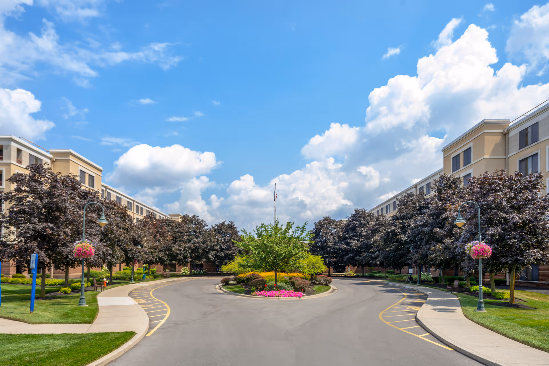 Driveway with a landscaped roundabout and hanging flower baskets leading to the front of a multi-story senior living building under a blue sky.