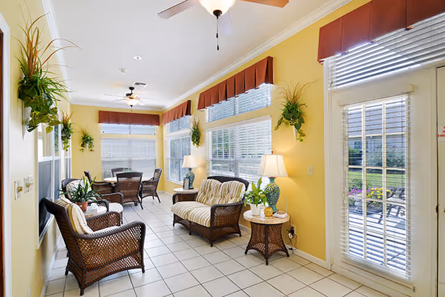 Bright and airy sunroom with yellow walls, white tiled floor, and large windows with red valances. The room is furnished with wicker chairs and a loveseat with cushions, a round wicker table with a lamp and plant, and a dining table with chairs near the windows. Several green plants hang on the walls and the room opens to an outdoor patio through a glass door with blinds.