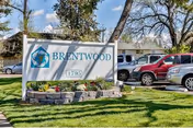 Outdoor view of the Brentwood Post-Acute facility sign with the address number 1795 displayed. The sign is surrounded by a small landscaped area with grass and plants. Several parked cars and trees are visible in the background under a partly cloudy sky.