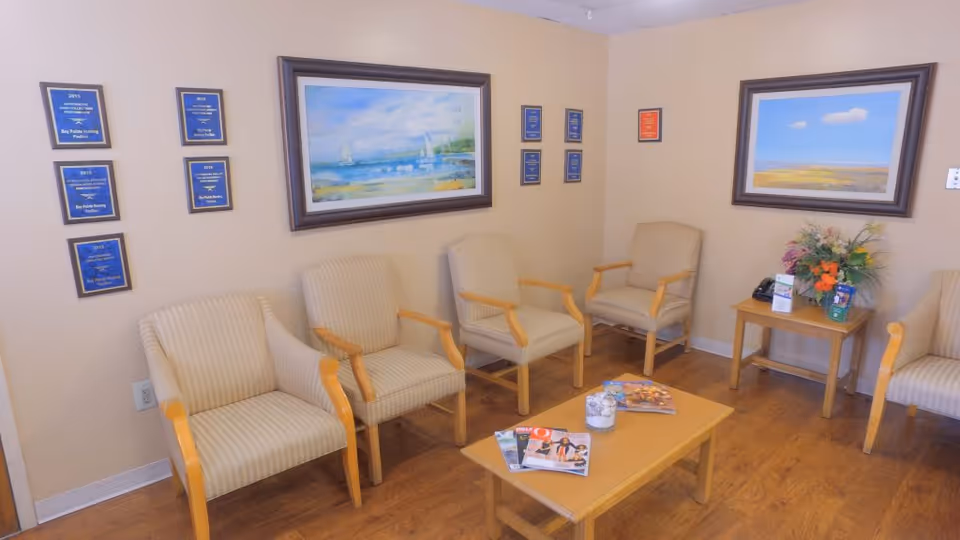 A waiting room with six beige armchairs arranged along two walls. A wooden coffee table in the center holds magazines and a small decorative item. The walls are decorated with framed paintings of landscapes and several plaques. A small side table with a flower arrangement and informational brochures is in the corner.