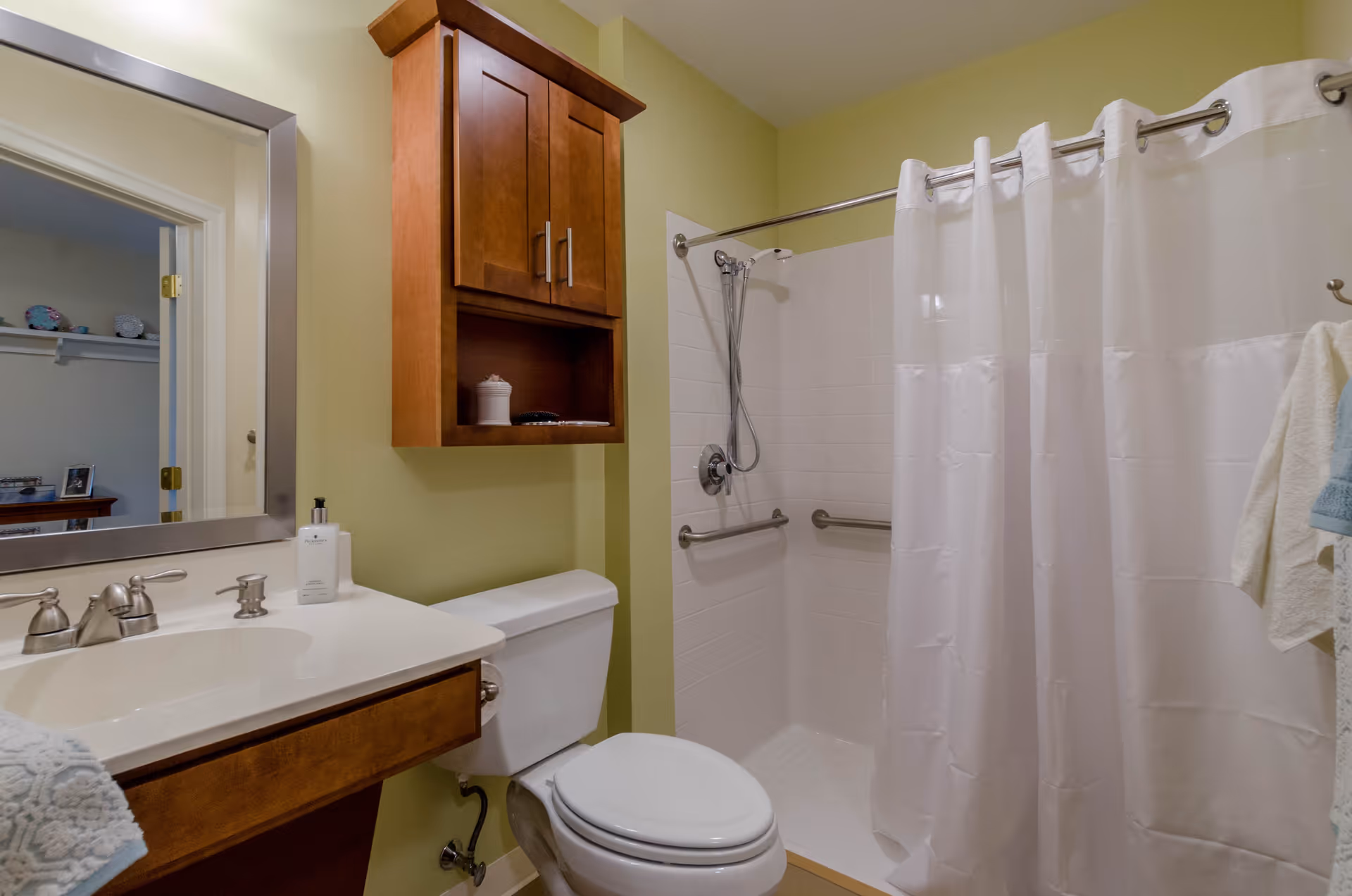 A bathroom with light green walls featuring a white toilet, a wooden cabinet mounted above the toilet, a white sink with a silver faucet, a large mirror, and a shower area with white tiled walls and a white shower curtain. There are grab bars installed inside the shower for safety.