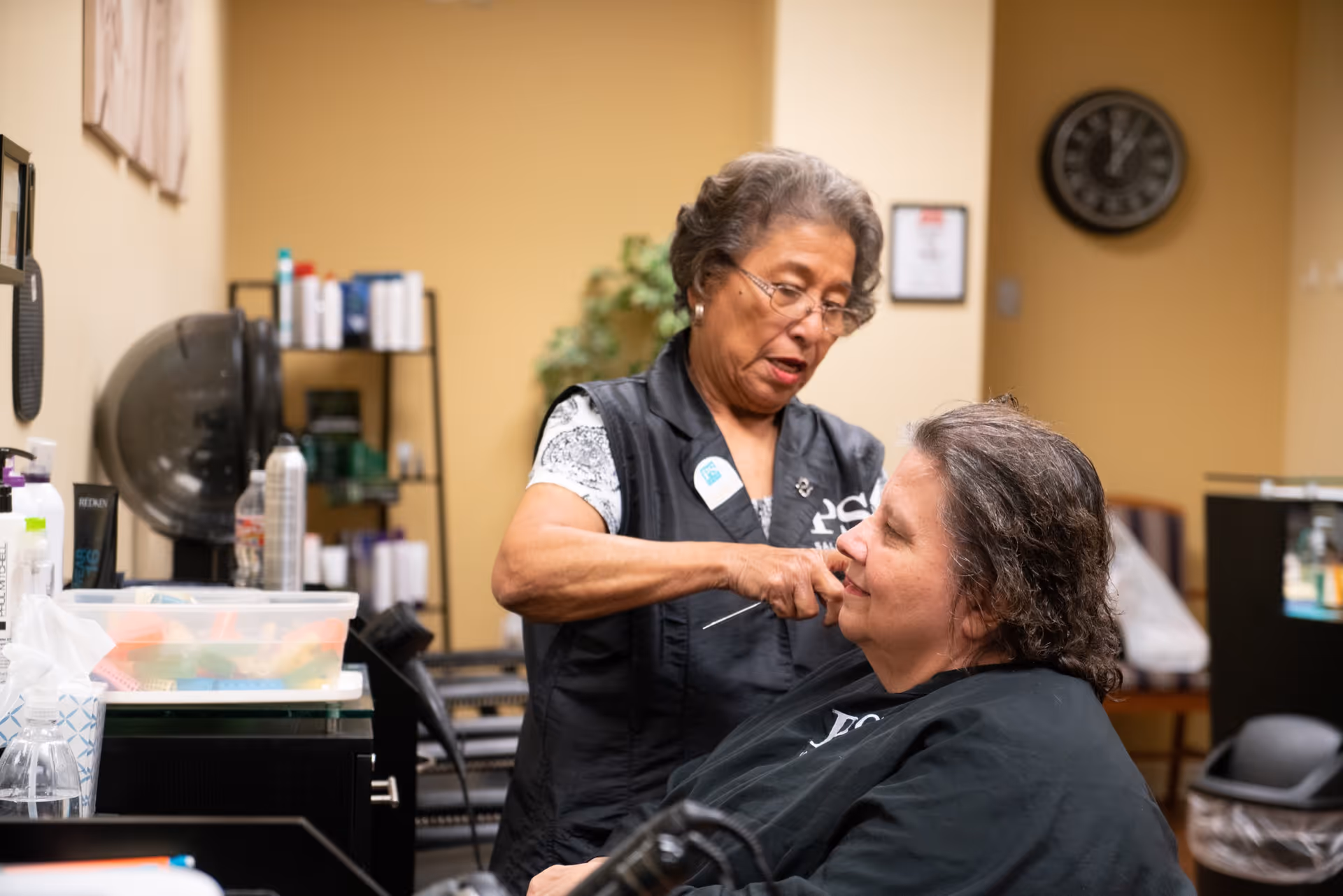 A hairstylist is cutting the hair of a seated woman in a salon setting. The hairstylist is focused on her work, and the woman receiving the haircut is smiling slightly. The room has shelves with hair products and a wall clock in the background.