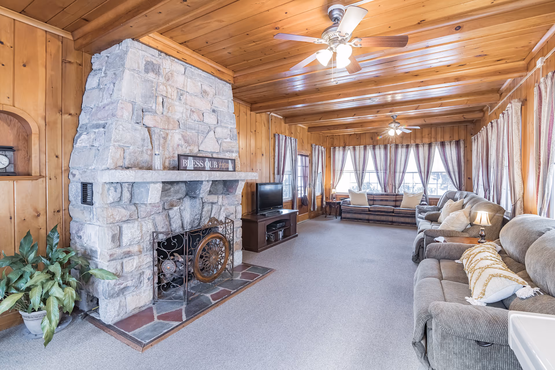 A spacious living room with wood-paneled walls and ceiling, featuring a large stone fireplace with a decorative screen. The room has multiple seating options including a gray sofa, armchairs, and a striped couch near large windows with sheer curtains. Ceiling fans with lights hang from the wooden ceiling, and a small TV sits on a stand next to the fireplace. A potted plant is placed near the fireplace.