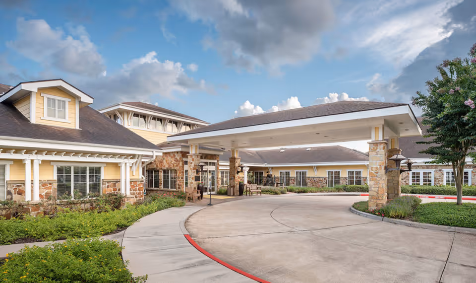 Front entrance with a covered porte-cochère of a yellow senior living building, curved driveway, and landscaping under a partly cloudy sky.
