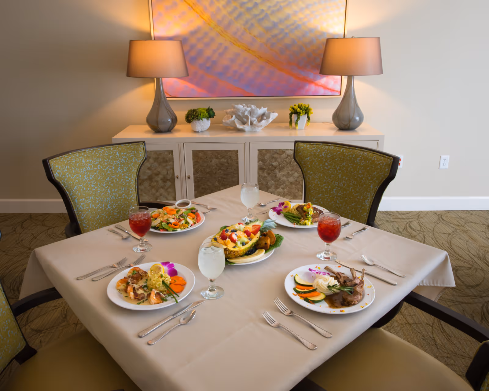 A dining table set for four with plated meals, glasses of beverages, and decorative chairs in a well-lit indoor dining room with a sideboard and lamps.