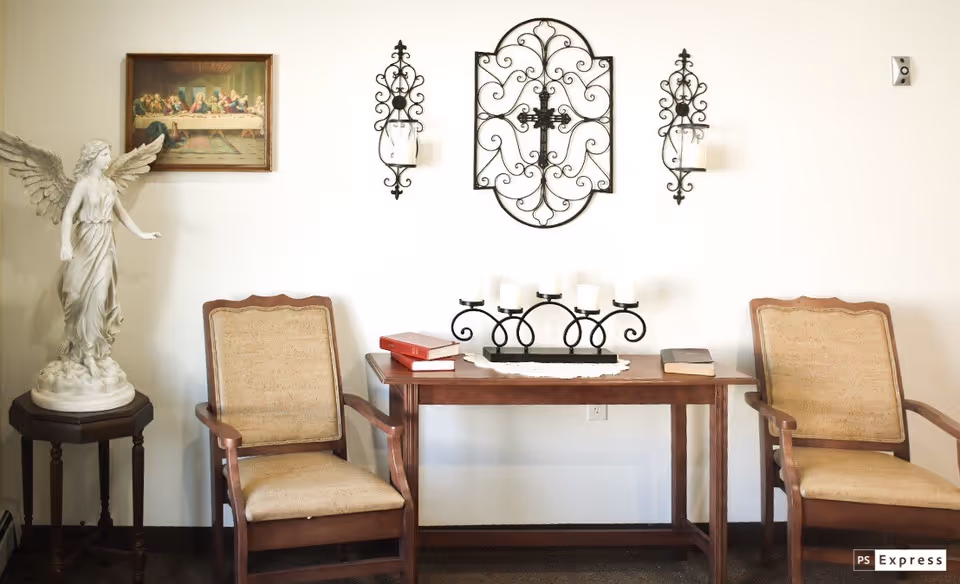 A quiet sitting area with two wooden armchairs with beige cushions on either side of a wooden table. On the table are three books and a decorative black candle holder with five white candles. Above the table on the wall is an ornate black metal wall decoration with a cross in the center, flanked by two matching candle sconces. To the left of the chairs is a small round table with a white angel statue. A framed picture of The Last Supper hangs on the wall above the left chair.