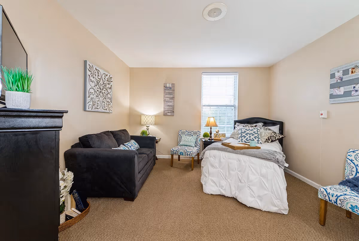 A cozy bedroom with a single bed covered in white bedding and multiple decorative pillows. Next to the bed is a small nightstand with a lamp and decorative items. Across from the bed is a dark gray sofa with patterned cushions. There are two patterned chairs, one near the window and one partially visible on the right. The walls are beige with framed artwork and a window with blinds letting in natural light. The floor is carpeted in a neutral tone.
