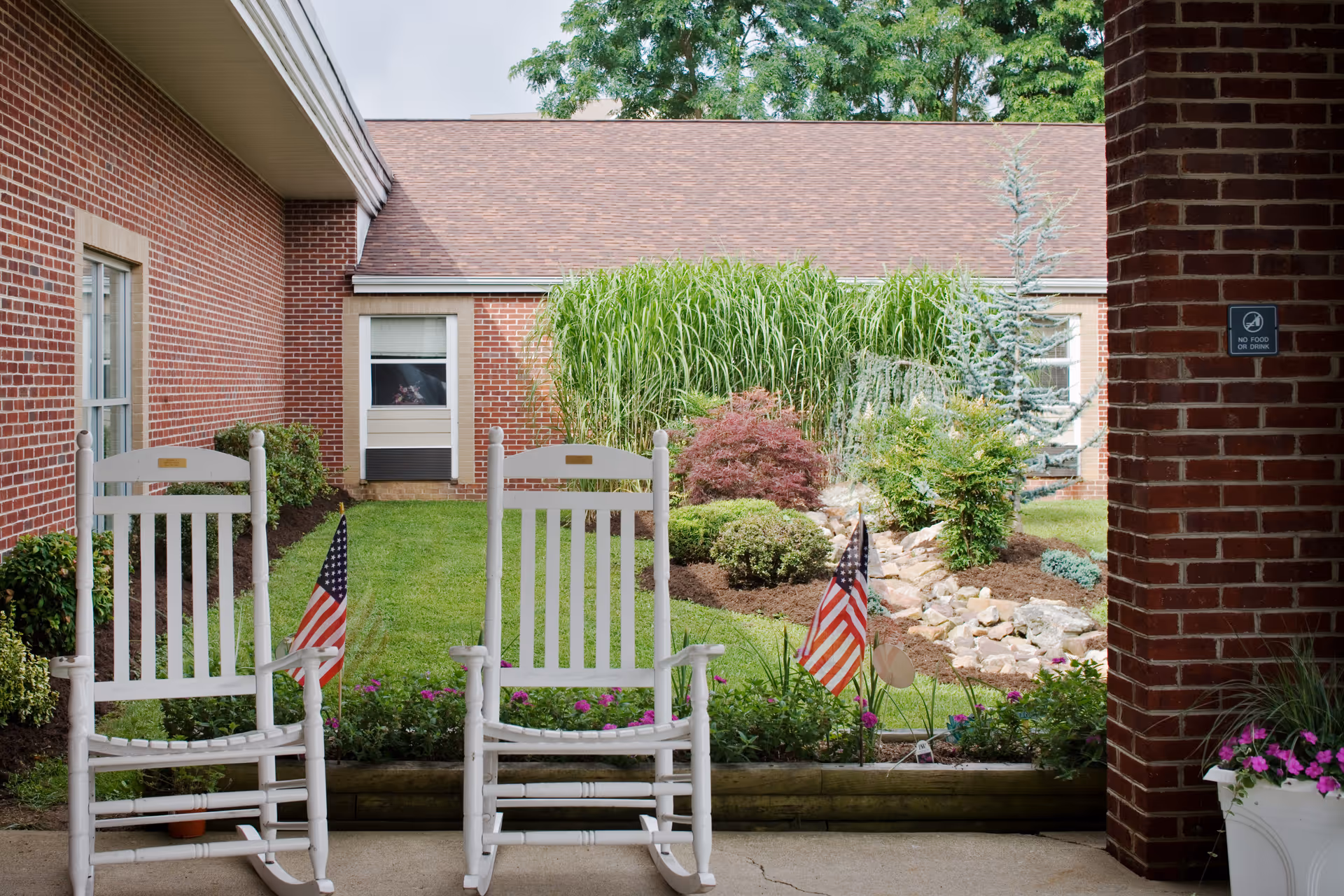 Two white rocking chairs with small American flags on a covered porch overlooking a landscaped courtyard and brick buildings.