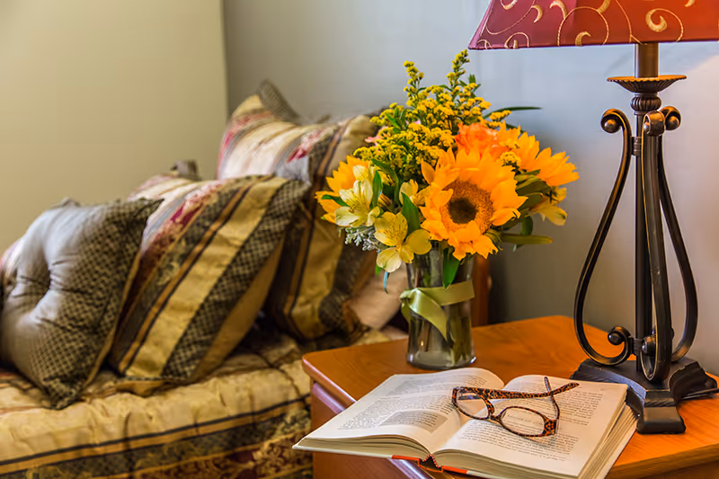 A cozy living room corner with a cushioned sofa adorned with patterned pillows, a wooden side table holding a vase of bright sunflowers and other flowers, an open book with reading glasses resting on it, and a decorative table lamp with a red patterned lampshade.