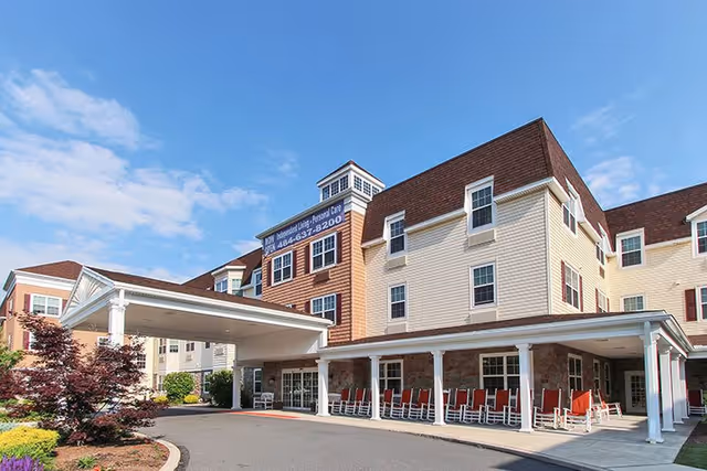 Exterior view of Keystone Villa at Fleetwood, a multi-story senior living facility with beige and brown siding, multiple windows, and a covered entrance with white pillars. There are red rocking chairs lined up under the covered porch area and landscaped greenery around the driveway.