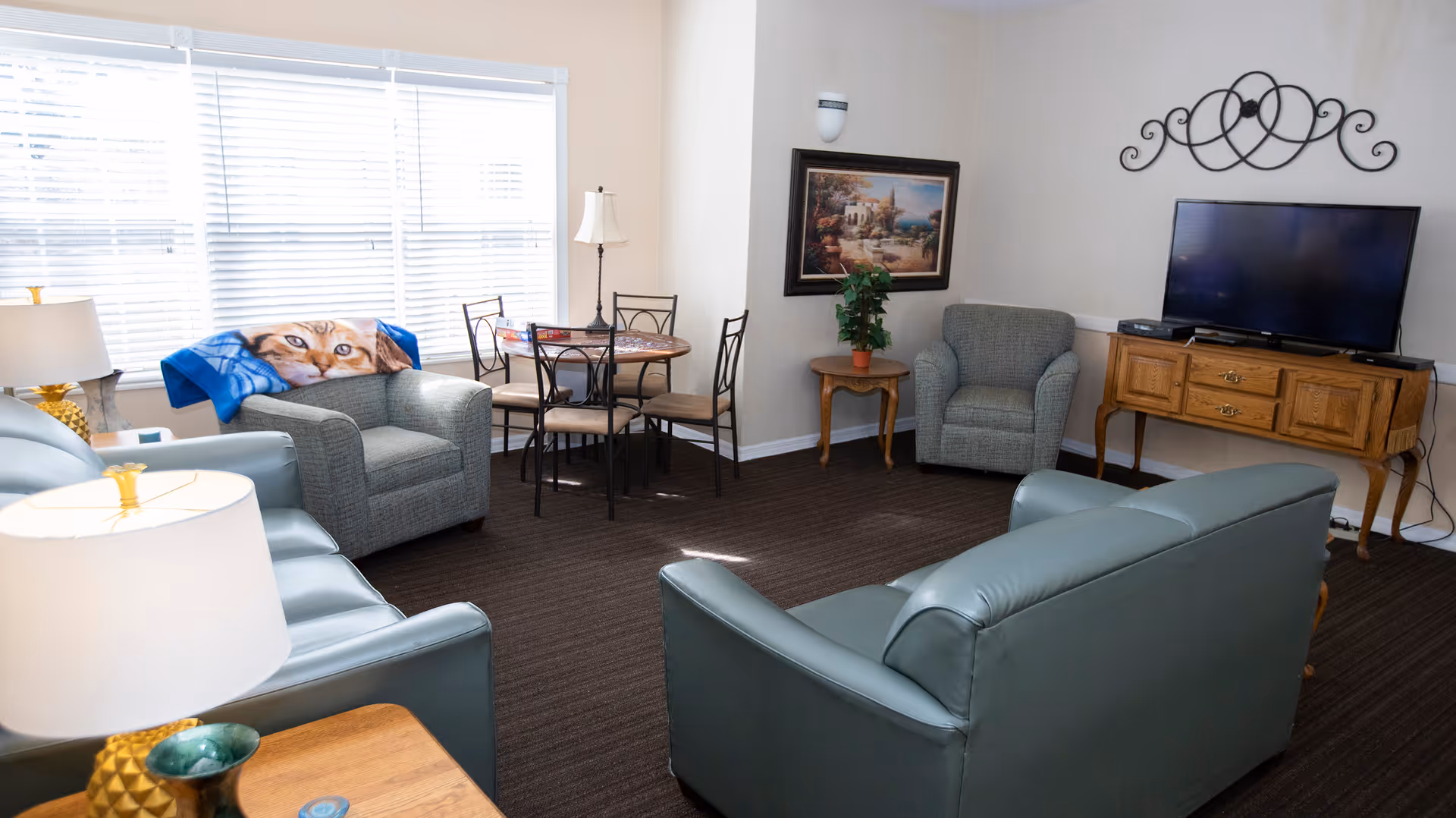Sunlit common living room with leather sofas and armchairs, a small dining table, and a TV on a wooden console.