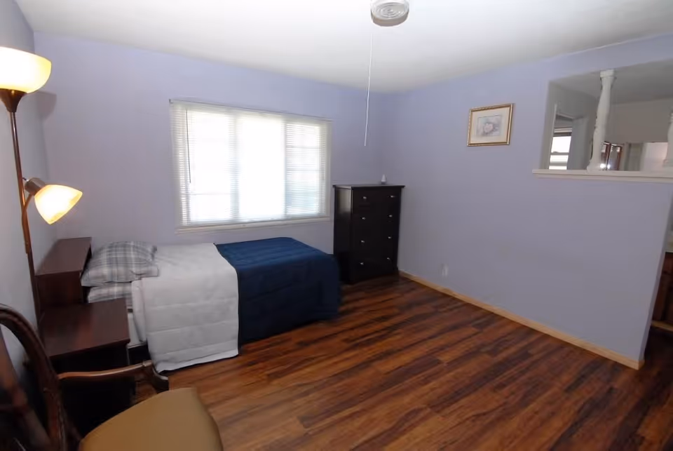 Small furnished bedroom with a single bed, window, dresser, floor lamp, and wood flooring.