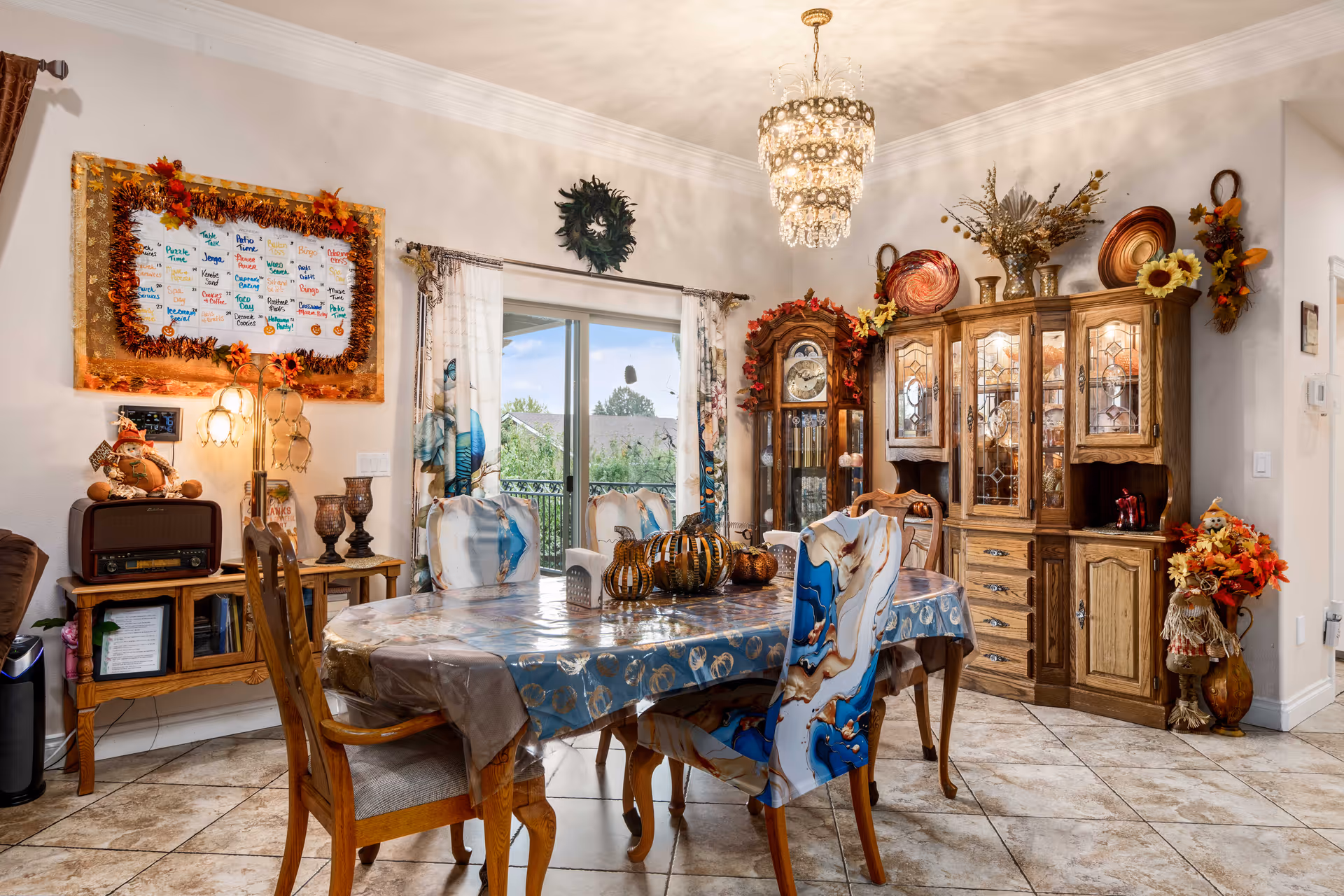 Dining room with a decorated table and chairs, a wooden china cabinet and grandfather clock, chandelier, and sliding glass doors to the outside.