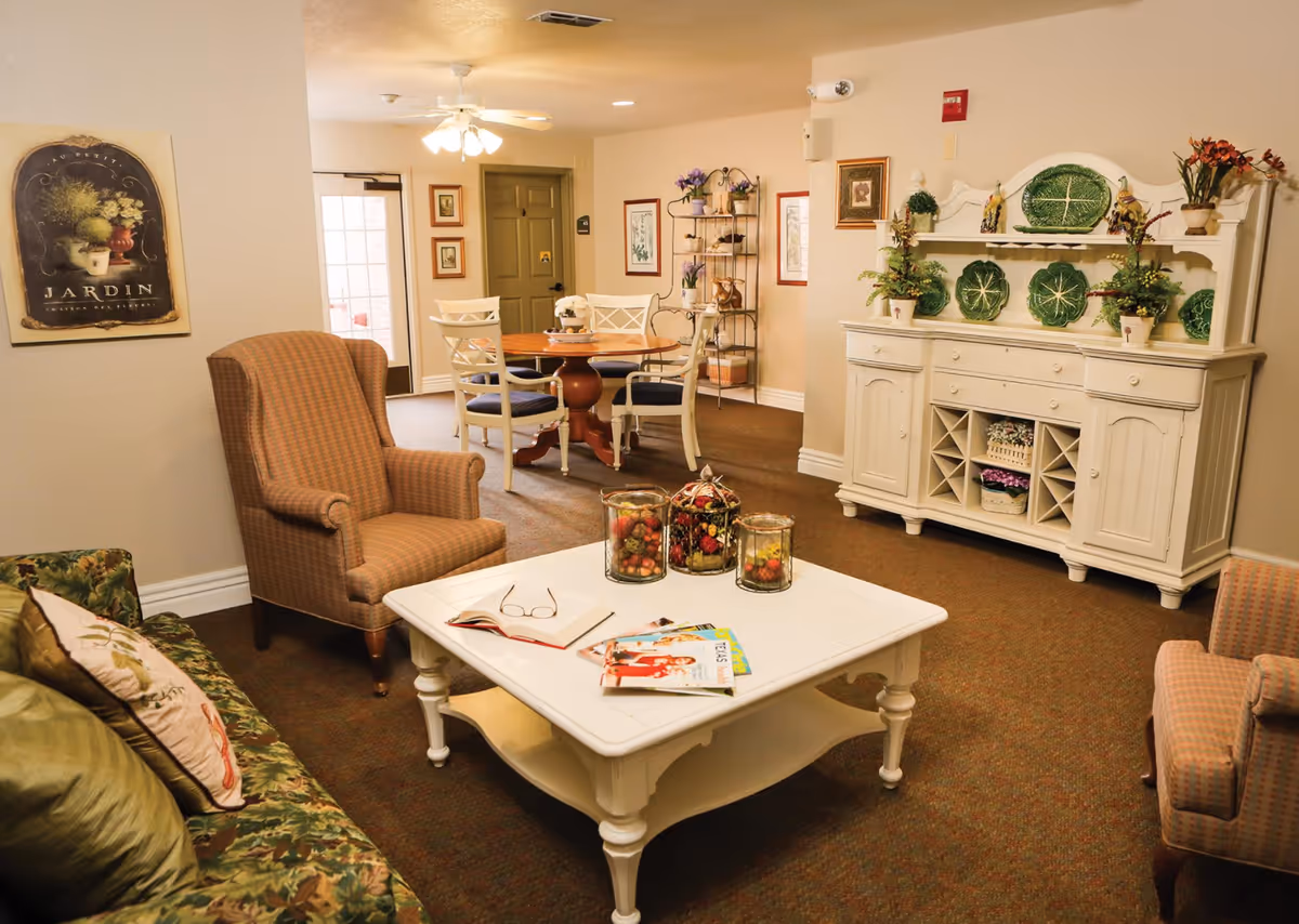 A cozy living room area in Bethesda Gardens Arlington Assisted Living featuring a floral patterned sofa with decorative pillows, two upholstered armchairs, a white wooden coffee table with magazines and decorative jars, a white sideboard with green decorative plates and plants, and a round wooden dining table with four white chairs in the background. The room is warmly lit with a ceiling fan light and decorated with framed artwork and plants.