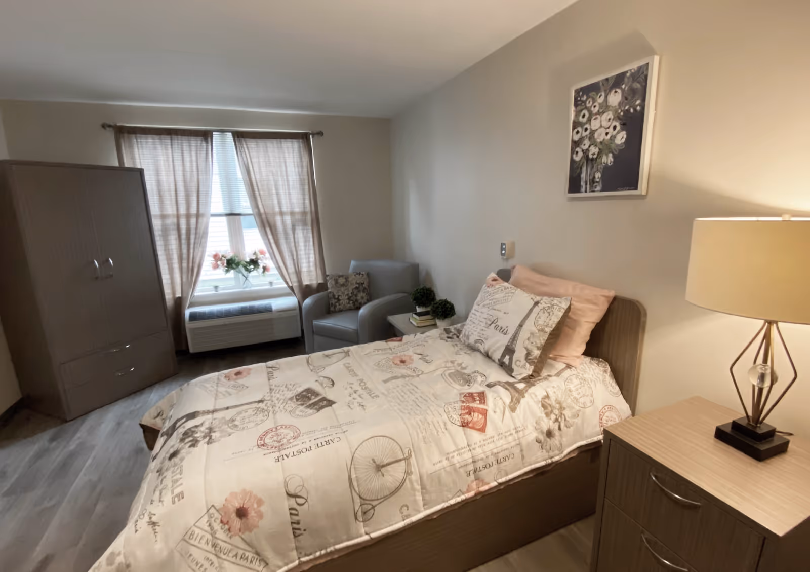 A cozy bedroom in a senior living facility featuring a single bed with Paris-themed bedding, a wooden nightstand with a modern lamp, a comfortable armchair with a decorative pillow, a small side table with two small potted plants and books, a window with beige curtains, and a wooden wardrobe.