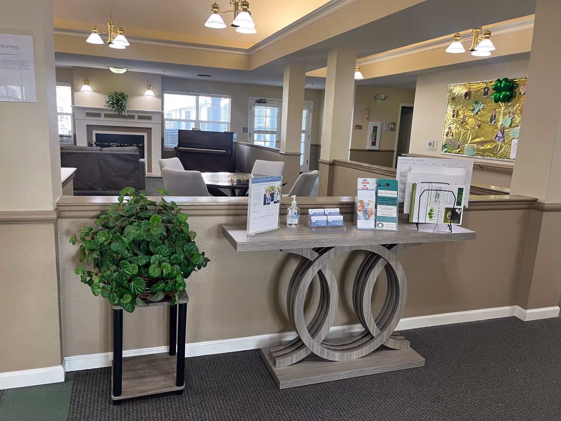 Interior view of a senior living facility common area with a decorative table holding brochures and hand sanitizer. A potted plant is on a stand to the left. In the background, there are chairs, a piano, a fireplace, and windows letting in natural light. The walls are beige with white trim, and ceiling lights illuminate the space.