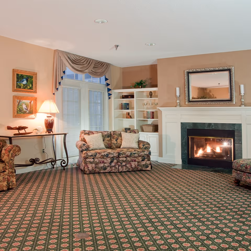 Cozy living room with patterned carpet, a floral loveseat, lit fireplace with mirror above, built-in bookshelves, and a lamp on a side table.