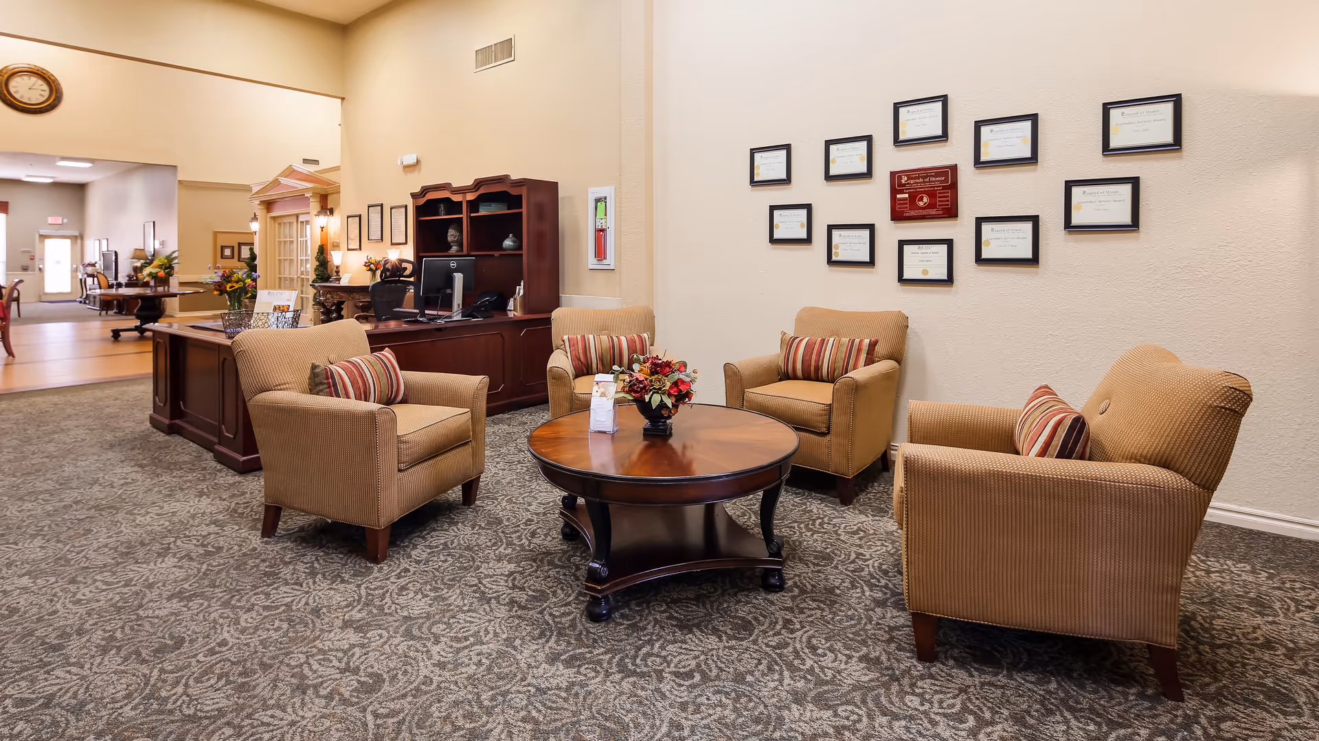 A cozy seating area in a senior living facility with four beige armchairs arranged around a round wooden coffee table. The chairs have striped cushions. Behind the seating area is a wall with multiple framed certificates and awards. To the left, there is a dark wooden reception desk with a computer and decorative items. The space has patterned carpet flooring and warm lighting.