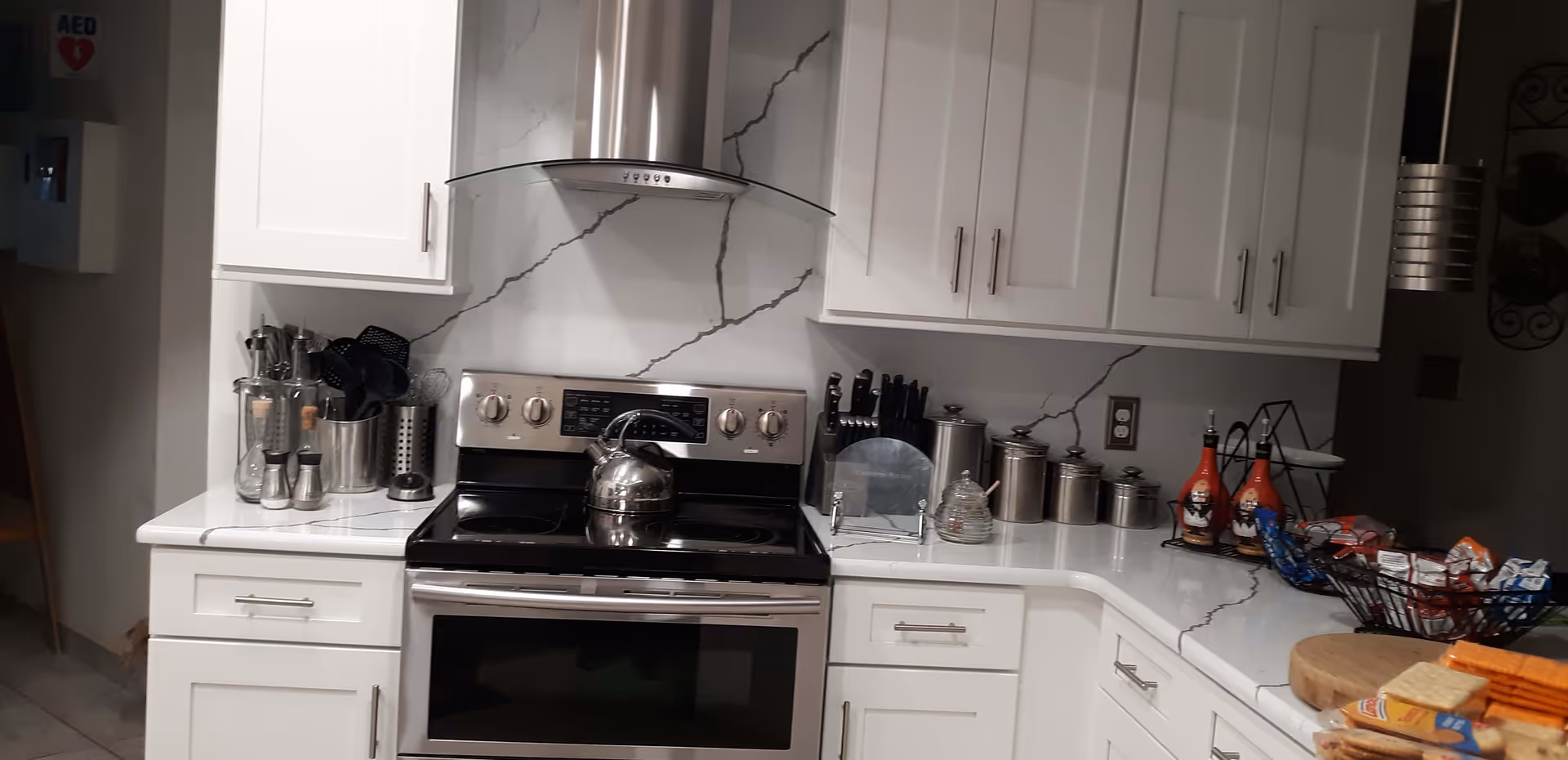 A modern kitchen with white cabinets and marble-patterned countertops and backsplash. There is a stainless steel stove with a kettle on it, a stainless steel range hood, various kitchen utensils in holders, a knife block, canisters, condiment bottles, and a basket of snacks on the counter.