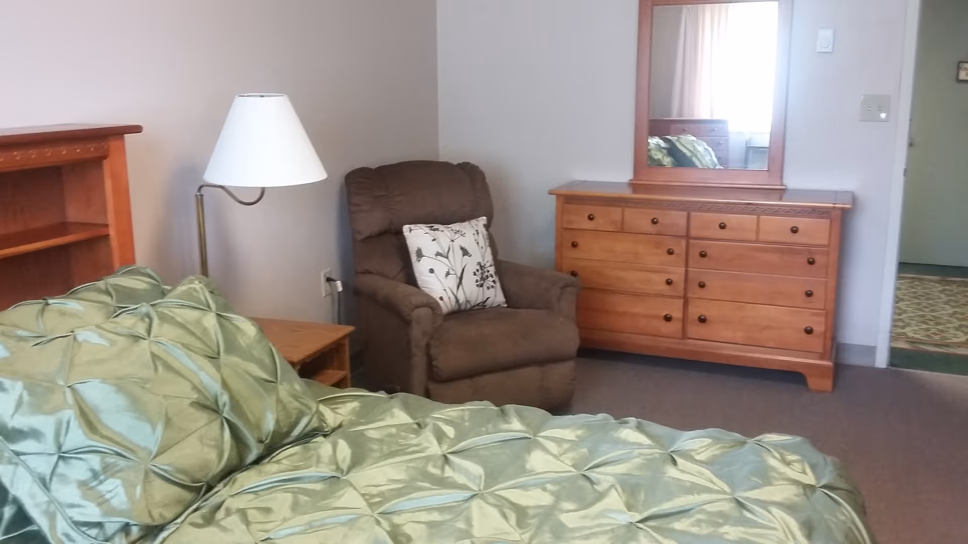 A bedroom with a green satin comforter on the bed, a wooden headboard, a brown recliner chair with a floral pillow, a wooden dresser with a mirror, and a floor lamp next to a small wooden side table.