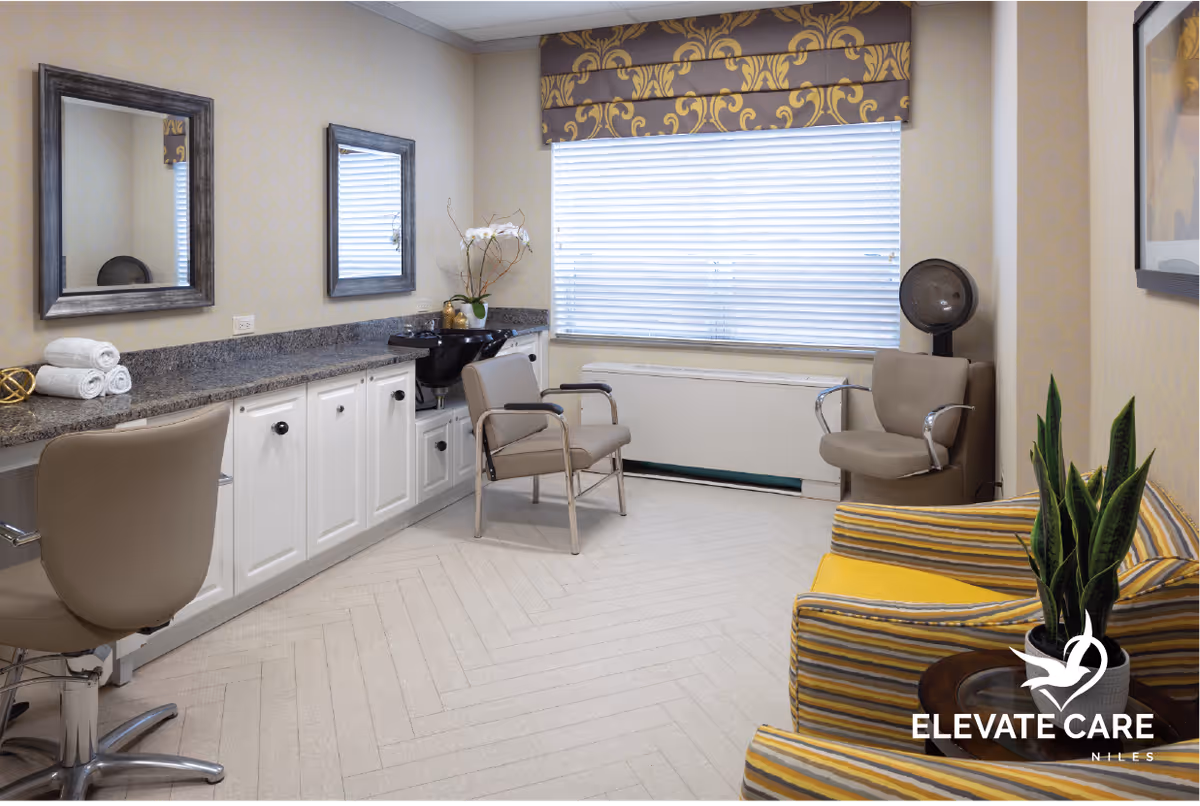 A well-lit salon area with beige walls and carpeted floor. The room features a long granite countertop with white cabinets underneath, two mirrors mounted on the wall, a black sink for hair washing, and three beige chairs including one salon chair with wheels. There is a window with blinds and a decorative valance, a potted plant on a small table, and a hair dryer chair in the corner. The room has a cozy and clean atmosphere.
