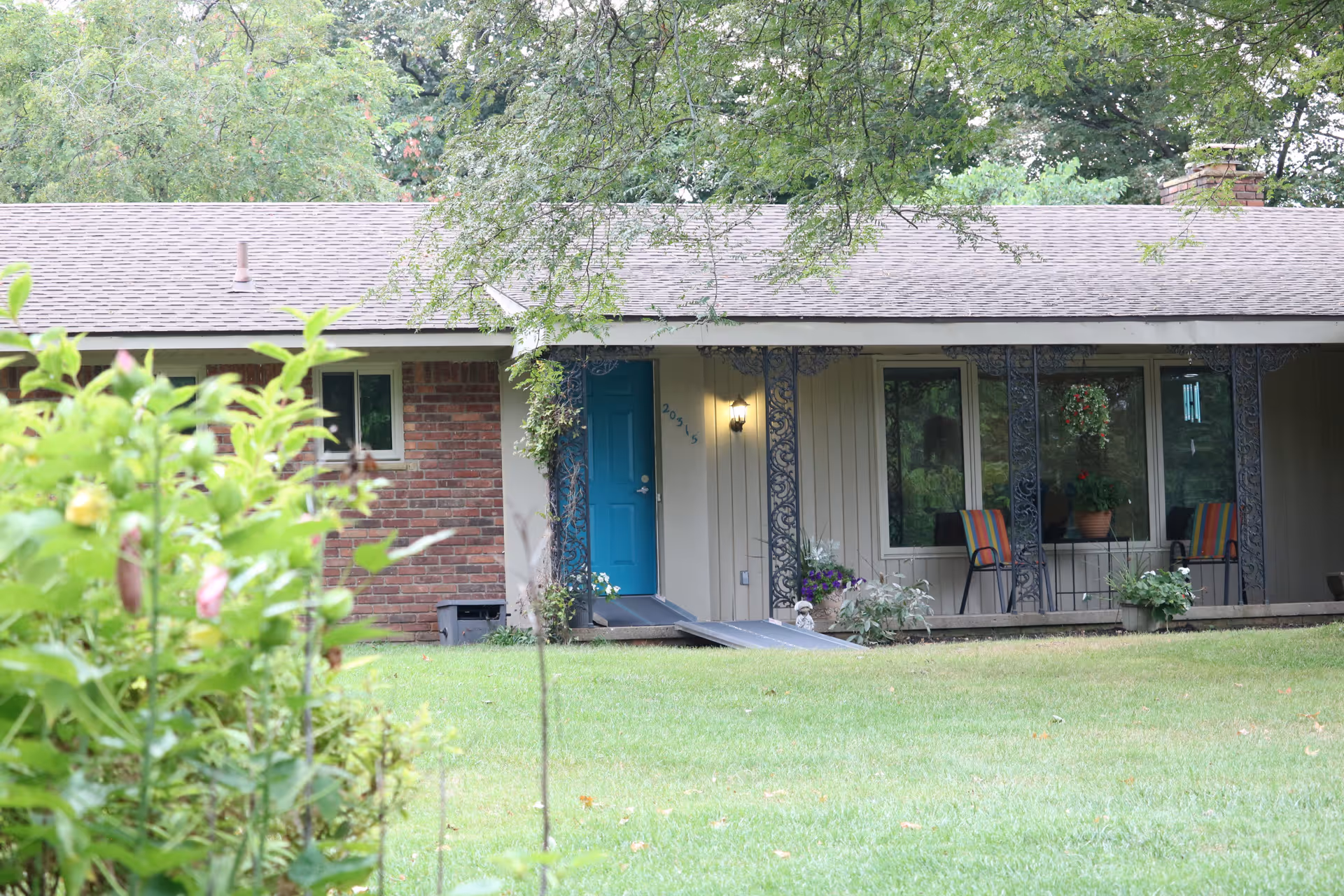 Front view of a single-story residential building with a blue door, brick and beige siding, a small porch with two chairs and potted plants, surrounded by green grass and trees.