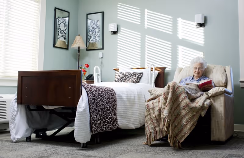 A sunlit retirement community bedroom with a made bed and a resident reading in a recliner under a blanket.