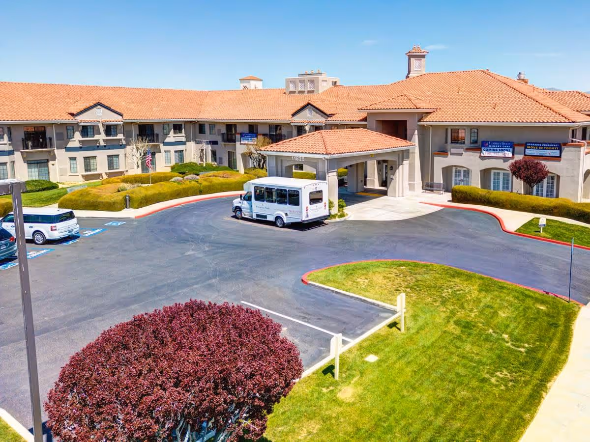 Exterior view of Whispering Winds of Apple Valley senior living facility showing a large building with a red-tiled roof, a driveway with a white shuttle bus parked under a covered entrance, several cars in parking spaces, well-maintained green lawns, and a purple-leaved tree in the foreground under a clear blue sky.