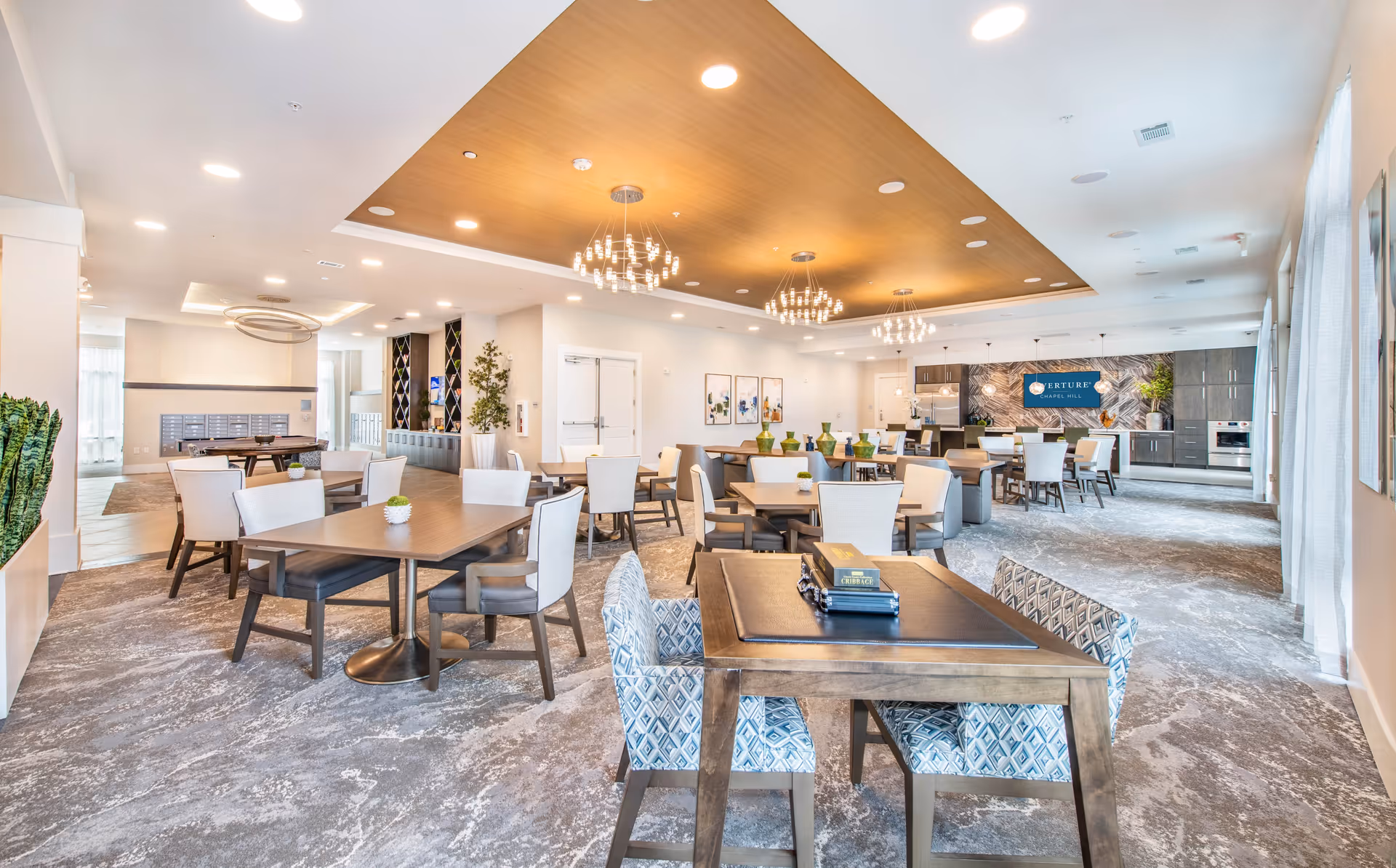 A spacious, well-lit common dining and activity area with multiple tables and chairs arranged throughout the room. The ceiling features modern chandeliers and recessed lighting with a wooden accent panel. The back wall has a kitchen area with dark cabinetry, a tiled backsplash, and a sign that reads 'Overture Chapel Hill'. The room has large windows with sheer curtains on the right side and decorative plants and artwork on the walls.