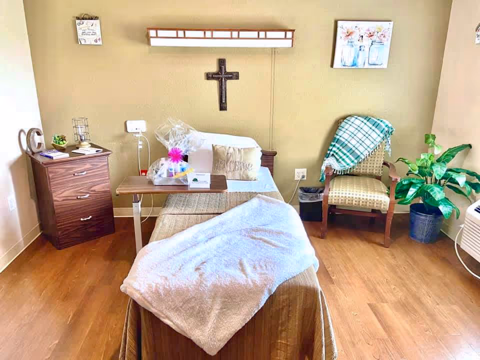A cozy nursing facility room with a single bed covered with a white blanket and a beige throw. There is a wooden bedside table with drawers on the left, holding a few decorative items and books. Above the bed on the wall is a wooden cross and a long light fixture. To the right of the bed is a cushioned chair with a green and white plaid blanket draped over it, next to a potted plant. The room has wooden flooring and light-colored walls with two framed pictures hanging.