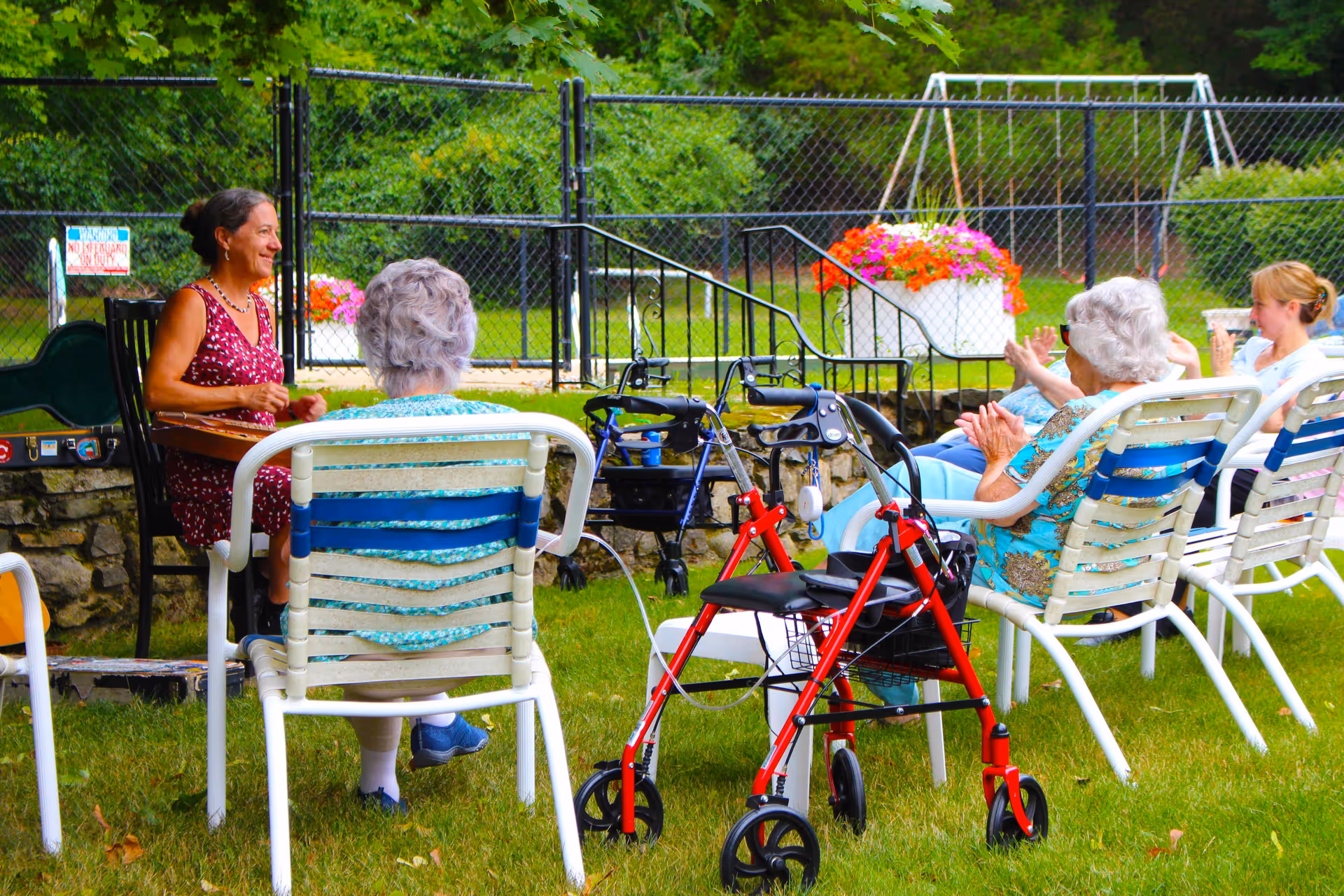 Several elderly residents sit on lawn chairs outdoors listening to a woman speaking, with walkers parked nearby on the grass.