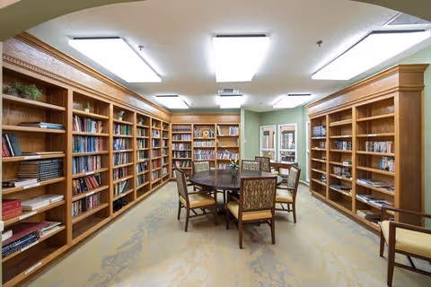 A well-lit library room with wooden bookshelves lining the walls filled with books. In the center, there is a round wooden table surrounded by six cushioned chairs. The room has a beige carpet and light green walls with a doorway leading to another area.