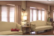 A cozy living room area with two beige sofas facing each other, a wooden side table with a lamp and decorative items, large windows with blinds and curtains, and a television in the corner.