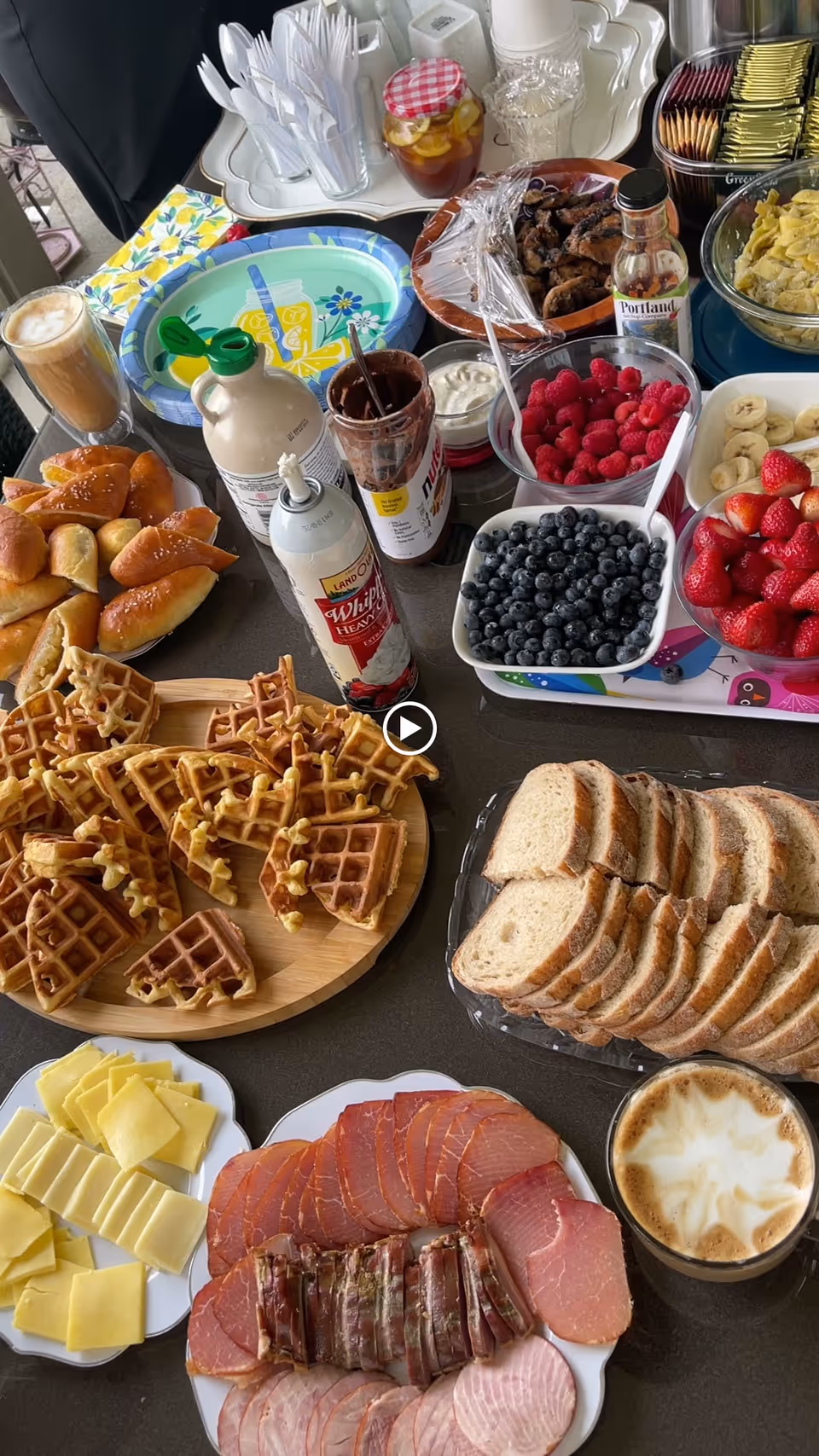 A table set with a variety of breakfast foods including waffles, sliced bread, assorted cheeses, cold cuts, fresh strawberries, blueberries, raspberries, sliced bananas, croissants, a cup of coffee with foam, whipped cream, syrup, and condiments. There are also plastic utensils and napkins in the background.
