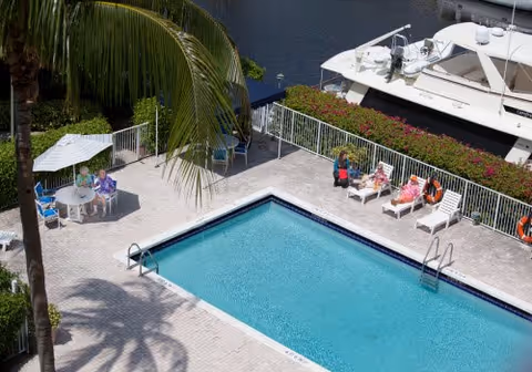 Rectangular outdoor swimming pool with lounge chairs and seated people near a table and umbrella, adjacent to a boat dock.
