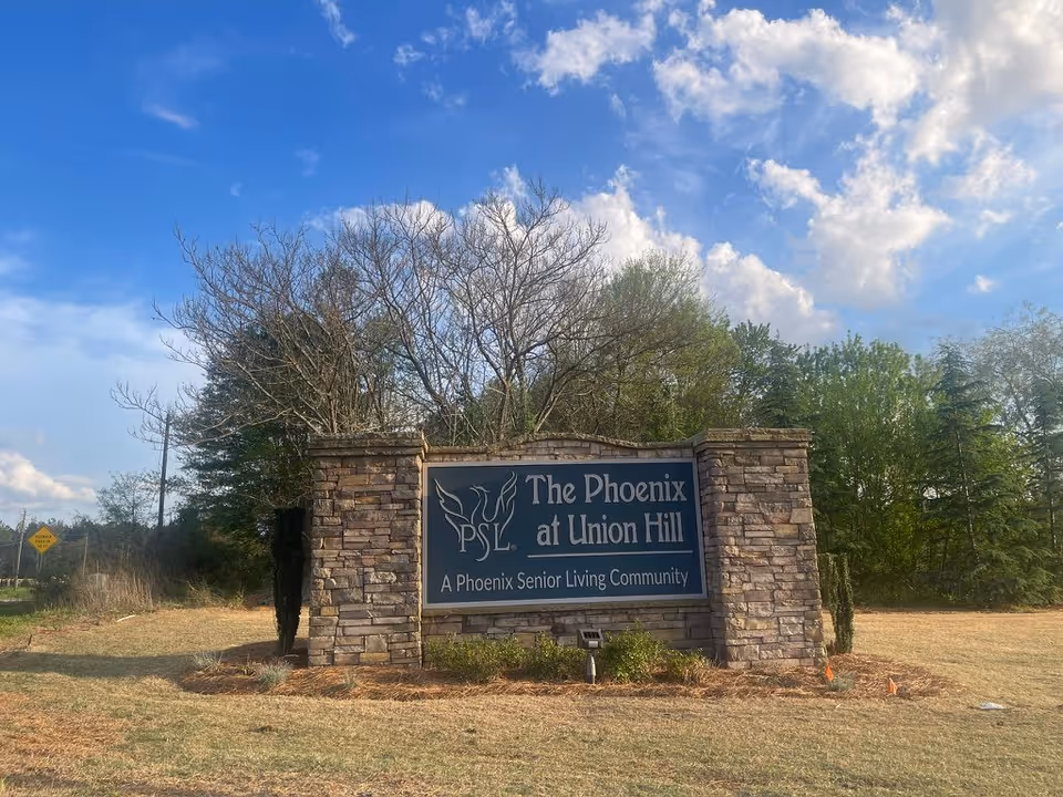 Stone entrance sign reading 'The Phoenix at Union Hill' for a senior living community set on a grassy lot with trees and a blue sky.