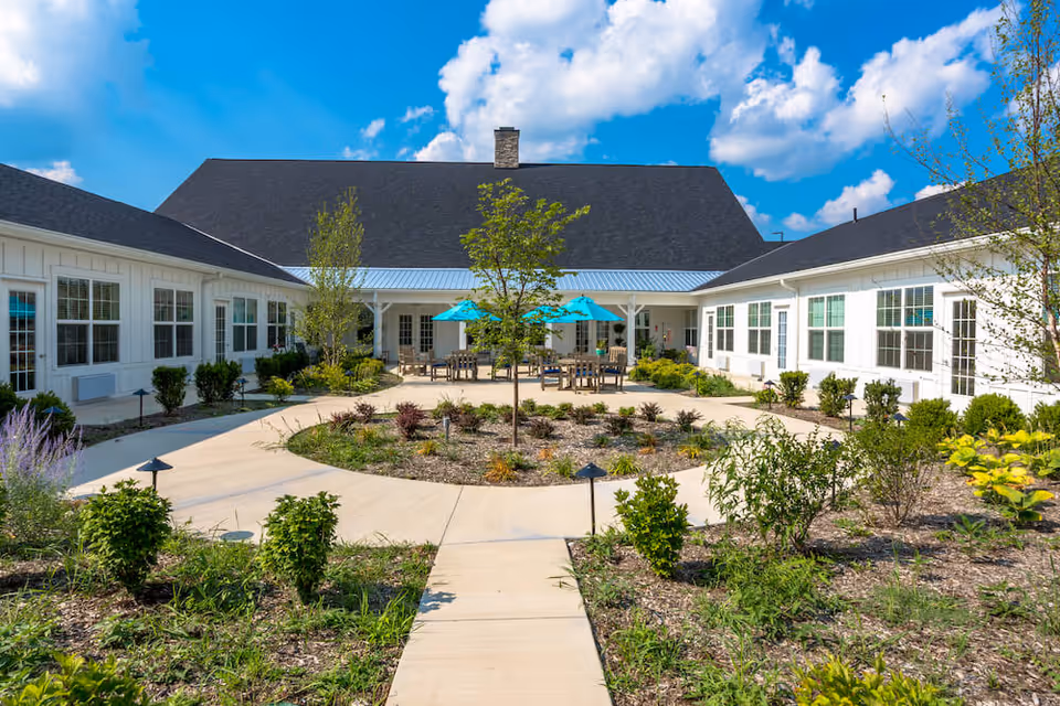 Outdoor courtyard area of a senior living facility with a circular walkway surrounding a landscaped garden. There are several tables with blue umbrellas and chairs under a covered patio attached to white buildings with many windows. The sky is blue with scattered clouds.