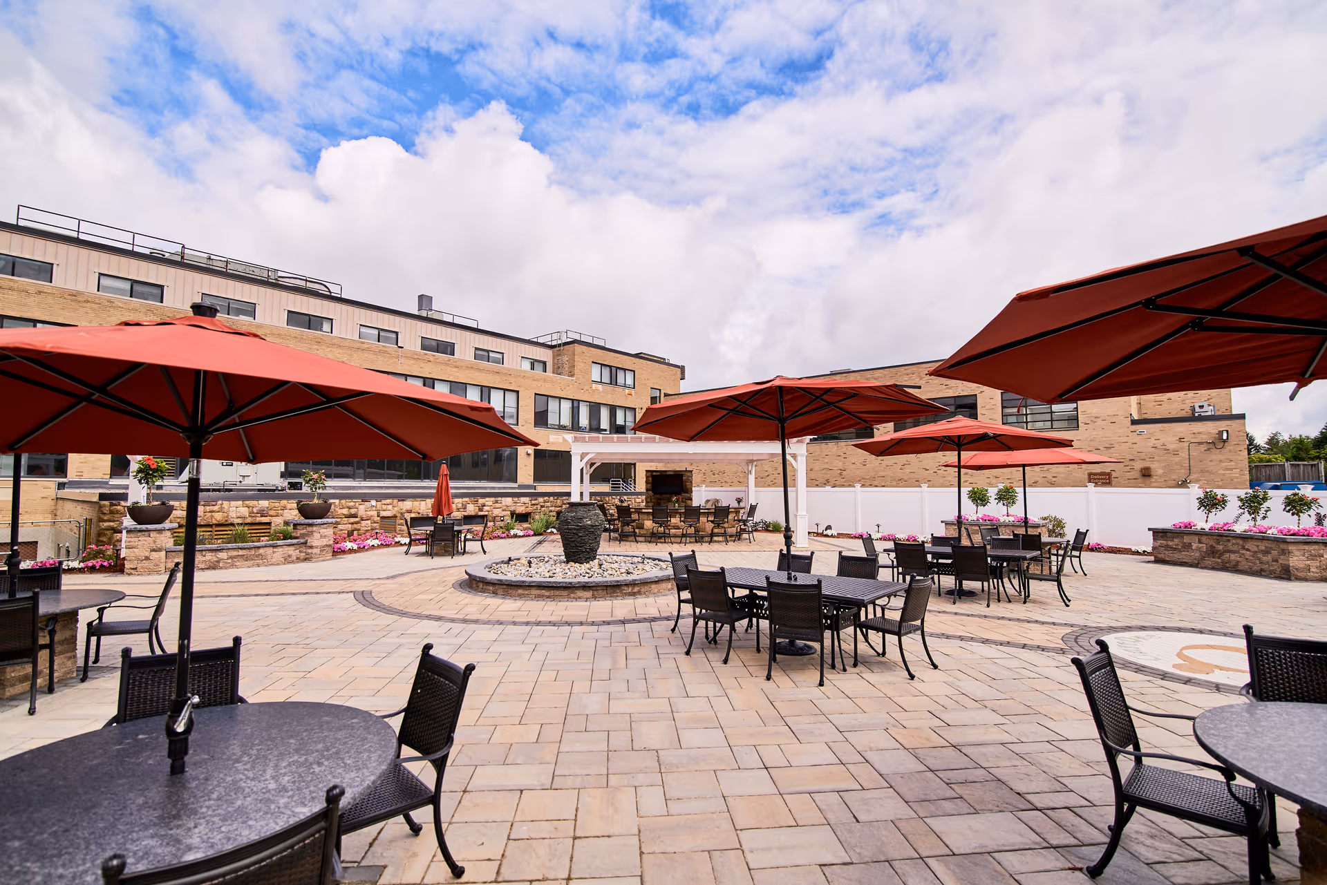 Outdoor patio area at CareOne at Concord featuring multiple round tables with black chairs and large red umbrellas. The patio is paved with stone tiles and has a central water feature surrounded by seating. The background shows a multi-story brick building under a partly cloudy sky.