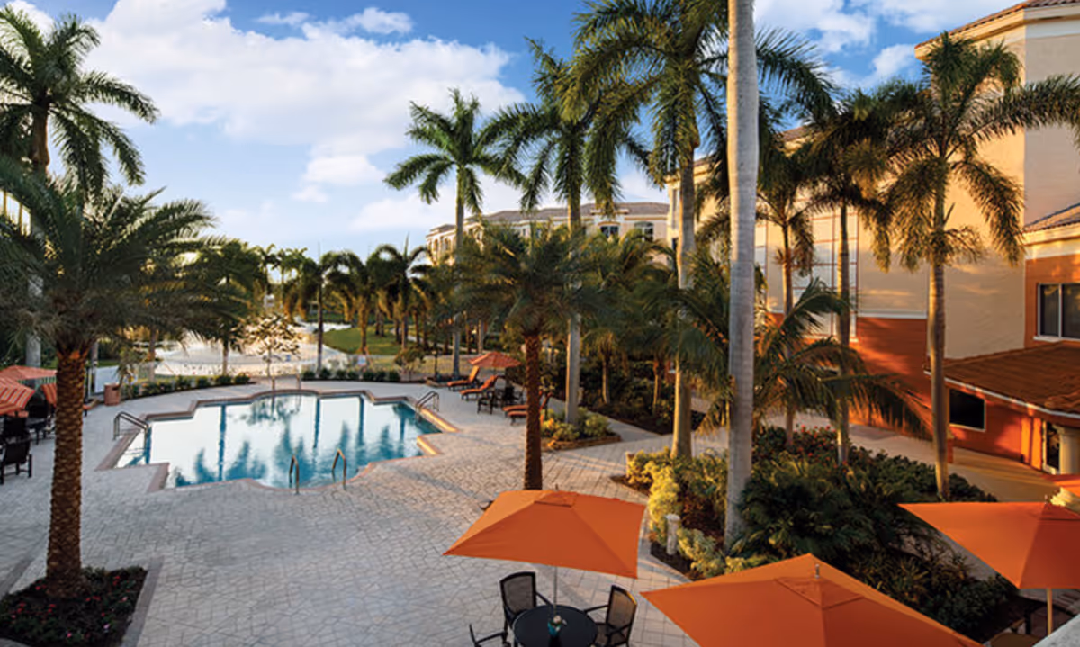 Outdoor area of Devonshire Senior Living Community at PGA National featuring a swimming pool surrounded by palm trees, lounge chairs, and tables with orange umbrellas under a partly cloudy sky.