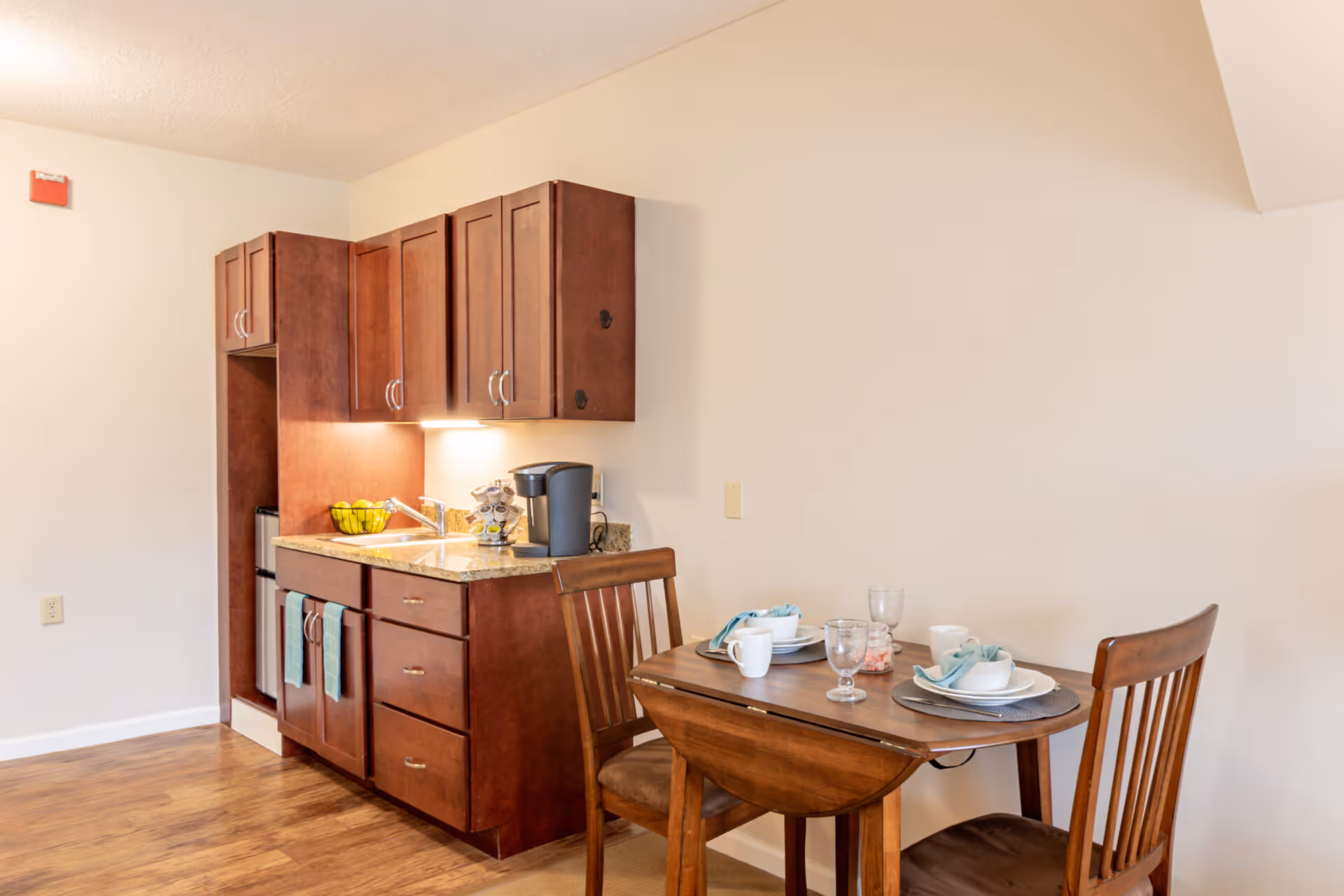 A small kitchen area with dark wood cabinets, a granite countertop, a sink, a coffee maker, and a bowl of green apples. Next to the kitchen is a wooden dining table set for two with plates, bowls, cups, and glasses. The room has light-colored walls and wood flooring.