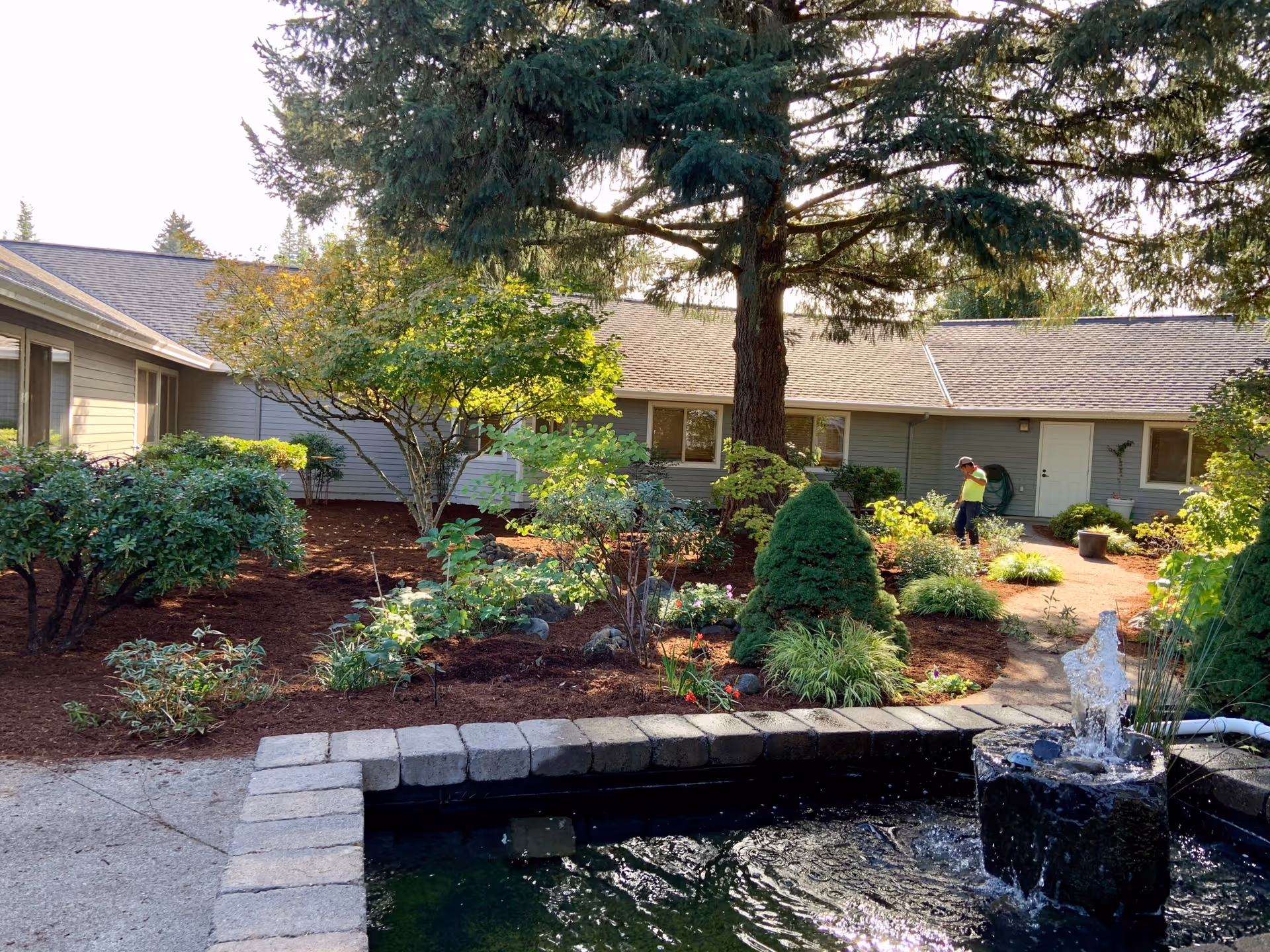 Landscaped courtyard with a small fountain, garden beds and a large tree in front of a single-story senior living building with a worker tending plants.