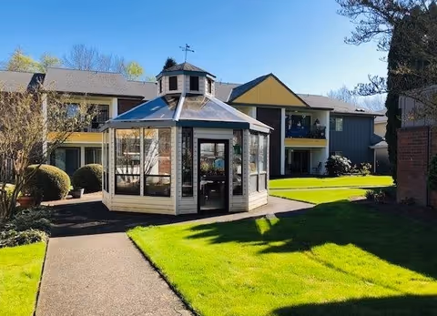 Outdoor view of a senior living facility courtyard with a glass octagonal gazebo in the center, surrounded by green grass, paved walkways, and a two-story building with balconies in the background under a clear blue sky.