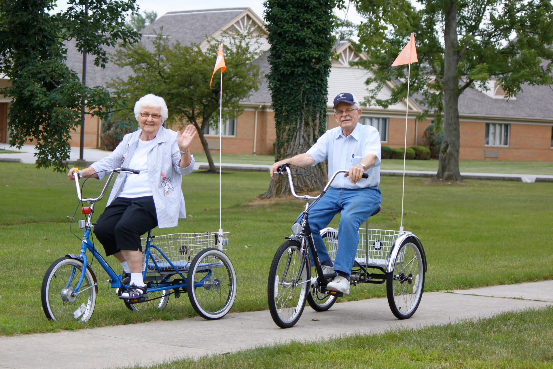 An elderly woman and man riding three-wheeled bicycles on a sidewalk in a grassy area with trees and a building in the background. The woman is waving and smiling, and both are dressed casually.