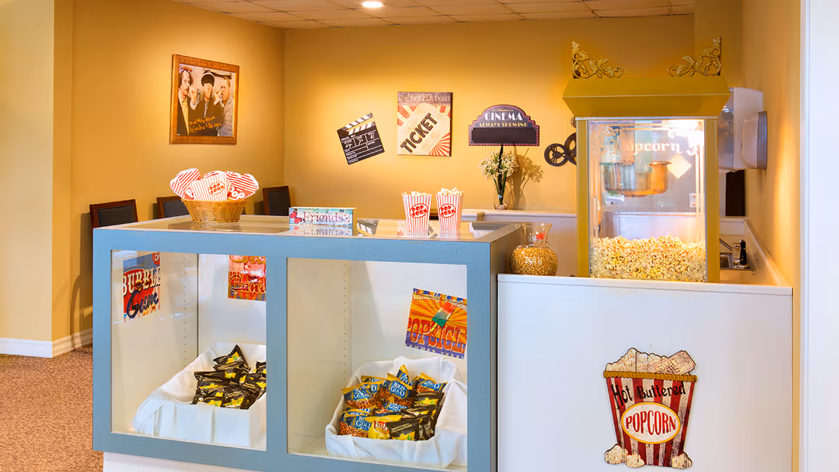A cozy popcorn and snack station in a senior living facility with a popcorn machine filled with freshly popped popcorn, baskets of popcorn bags, and bins of assorted snacks. The wall behind features vintage-style movie-themed decorations including a clapperboard, ticket, and cinema sign. There are chairs and a framed photo on the adjacent wall.