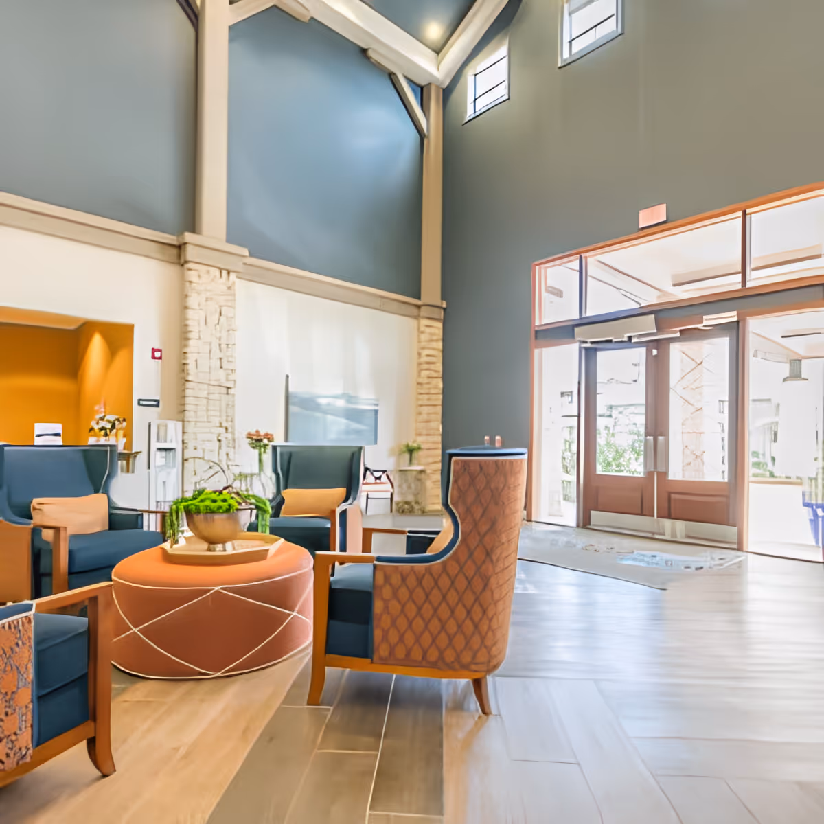 Bright high-ceilinged lobby with seating area of armchairs and an ottoman near large glass entrance doors.
