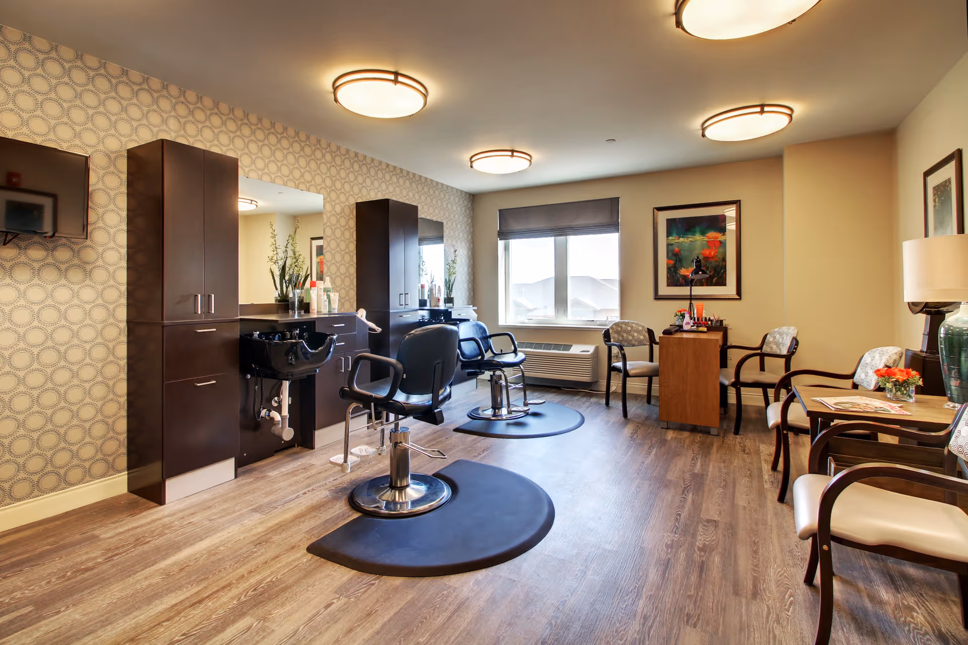 Interior of a senior living facility hair salon with two black salon chairs on black mats in front of mirrors and dark wood cabinetry. The room has patterned wallpaper on one wall, wood flooring, a window with a view outside, and several chairs arranged around small tables with flowers and magazines.