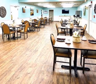 A spacious dining room with multiple tables and chairs arranged neatly. The room has light blue walls, wood flooring, and several framed pictures on the walls. There is a television mounted on the far wall and a counter area with cabinets. The tables are set with cups, plates, and decorative centerpieces.