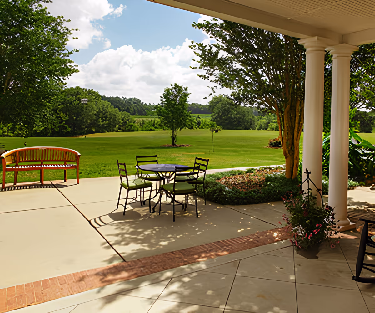 Outdoor patio area with a round table and four chairs with green cushions, a wooden bench, and large white columns. The patio overlooks a large grassy lawn with trees and a partly cloudy sky.