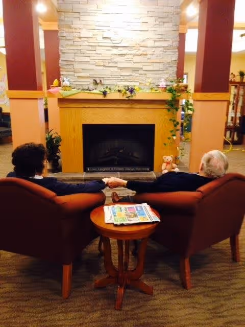 Two elderly people sitting in armchairs facing a stone fireplace in a cozy room, holding hands. A small wooden table with newspapers is between them. The room has warm lighting and decorative plants around the fireplace.