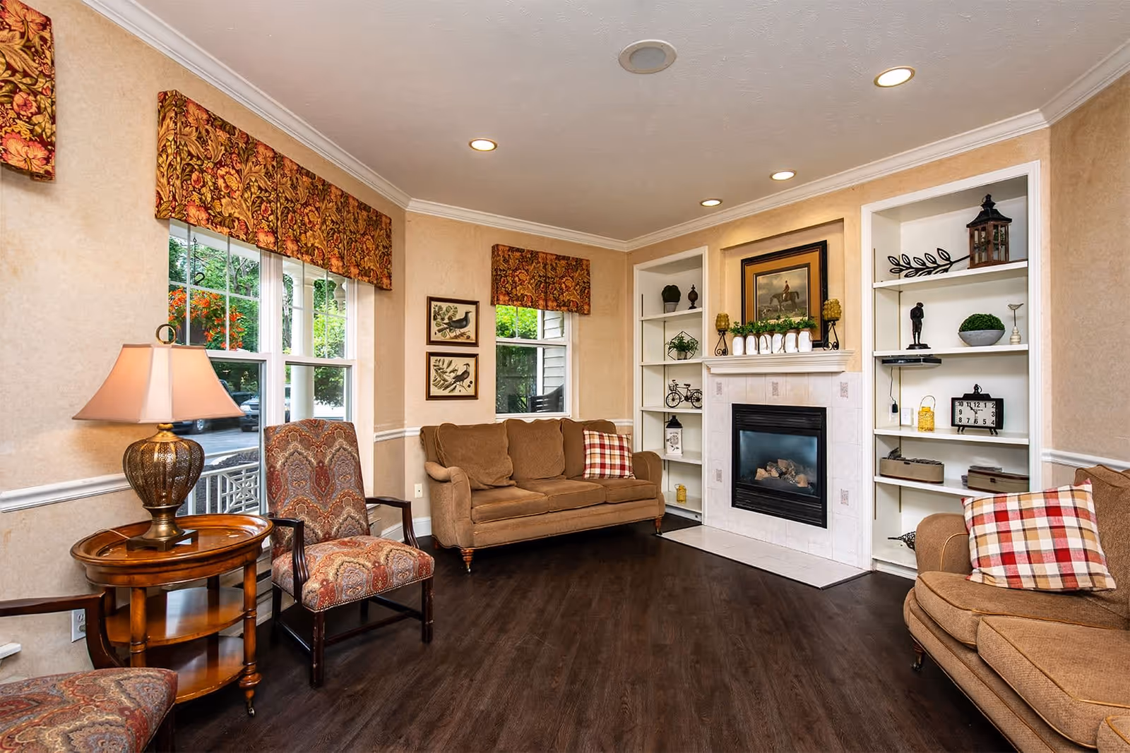 A cozy living room with two brown sofas, two patterned armchairs, a wooden side table with a lamp, and a fireplace surrounded by built-in white shelves decorated with various ornaments. The room has dark wood flooring, beige walls, and windows with floral valances letting in natural light.